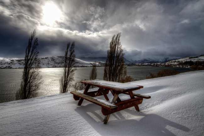 Обои картинки фото природа, зима, вода, скамейка, солнце, река, столик, деревья, снег, горы, тучи